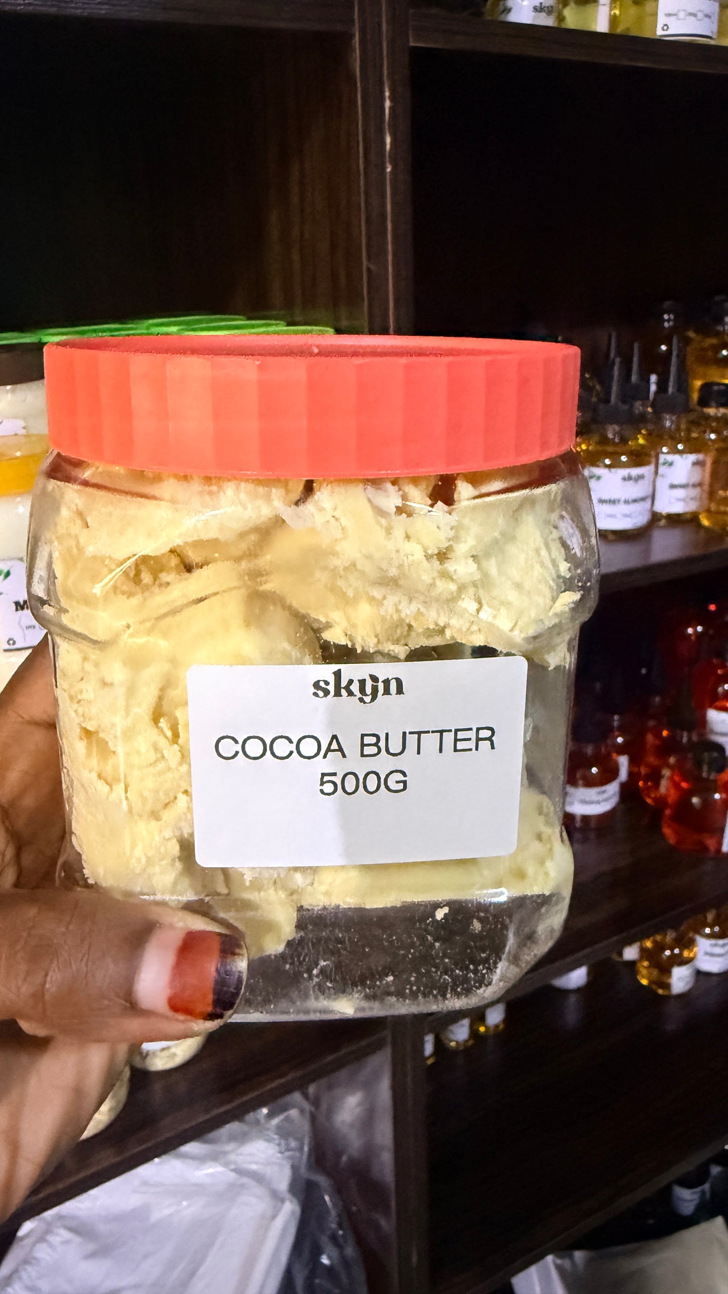 A hand holding a clear plastic jar of cocoa butter with a bright orange lid and a white label reading 'skyn COCOA BUTTER 500G'. The background shows dark wooden shelves stocked with other jars of ingredients or products.