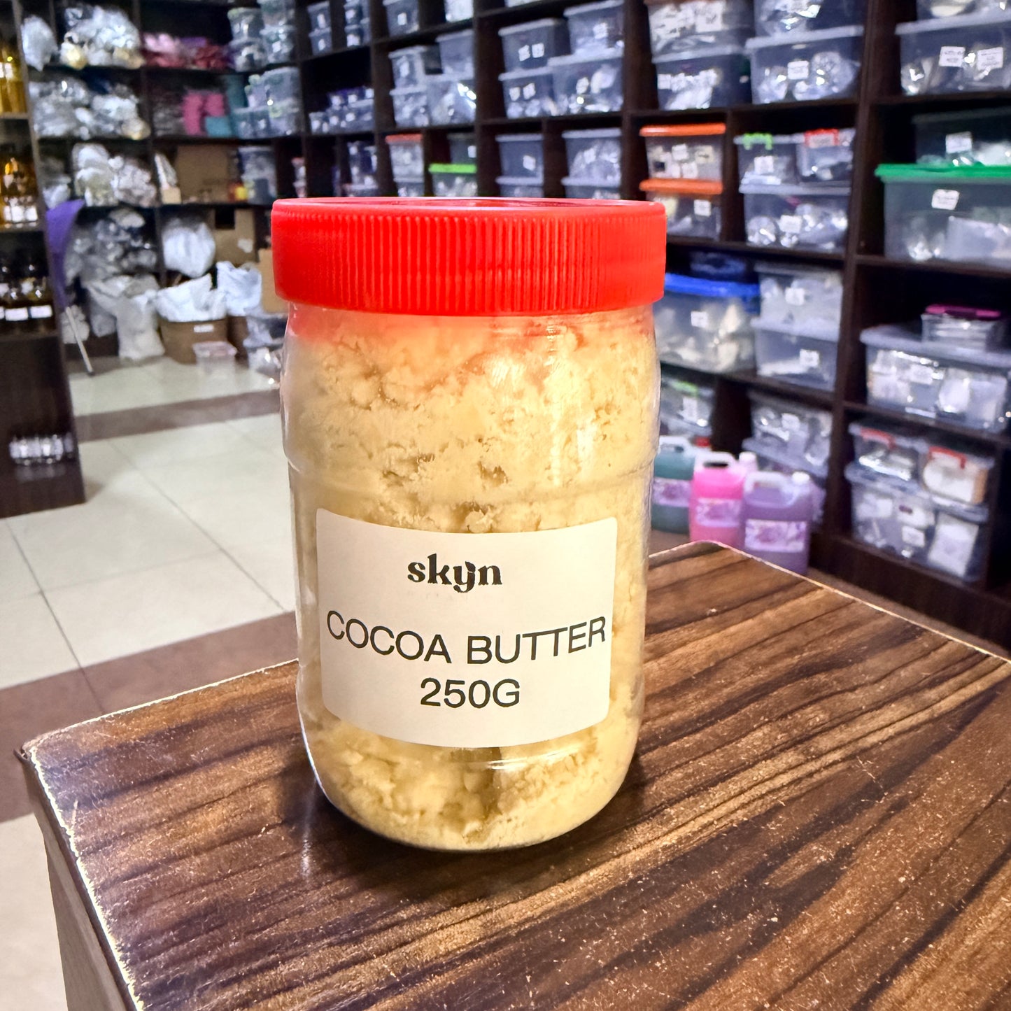 A 250g jar of skyn Cocoa Butter with a red lid and white label, sitting on a wooden table in a store setting with shelves of packaged goods in the background.