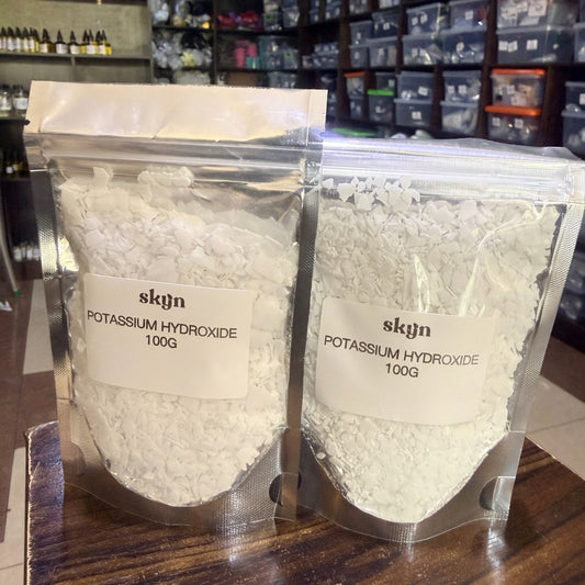 Two clear plastic bags filled with white potassium hydroxide powder, labeled 'skyn POTASSIUM HYDROXIDE 100G', displayed on a wooden surface with a storage room in the background.