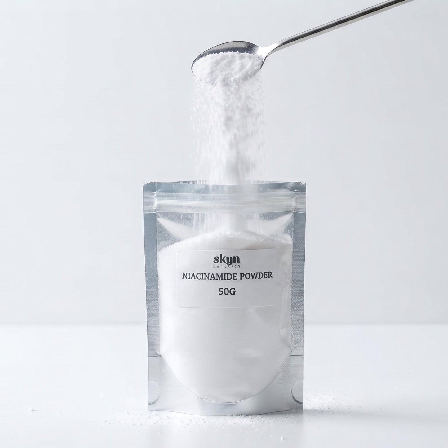 A silver stand-up pouch labeled 'skyn NIACINAMIDE POWDER 50G' on a white surface, with white powder being poured into it from a metal spoon against a light gray background.