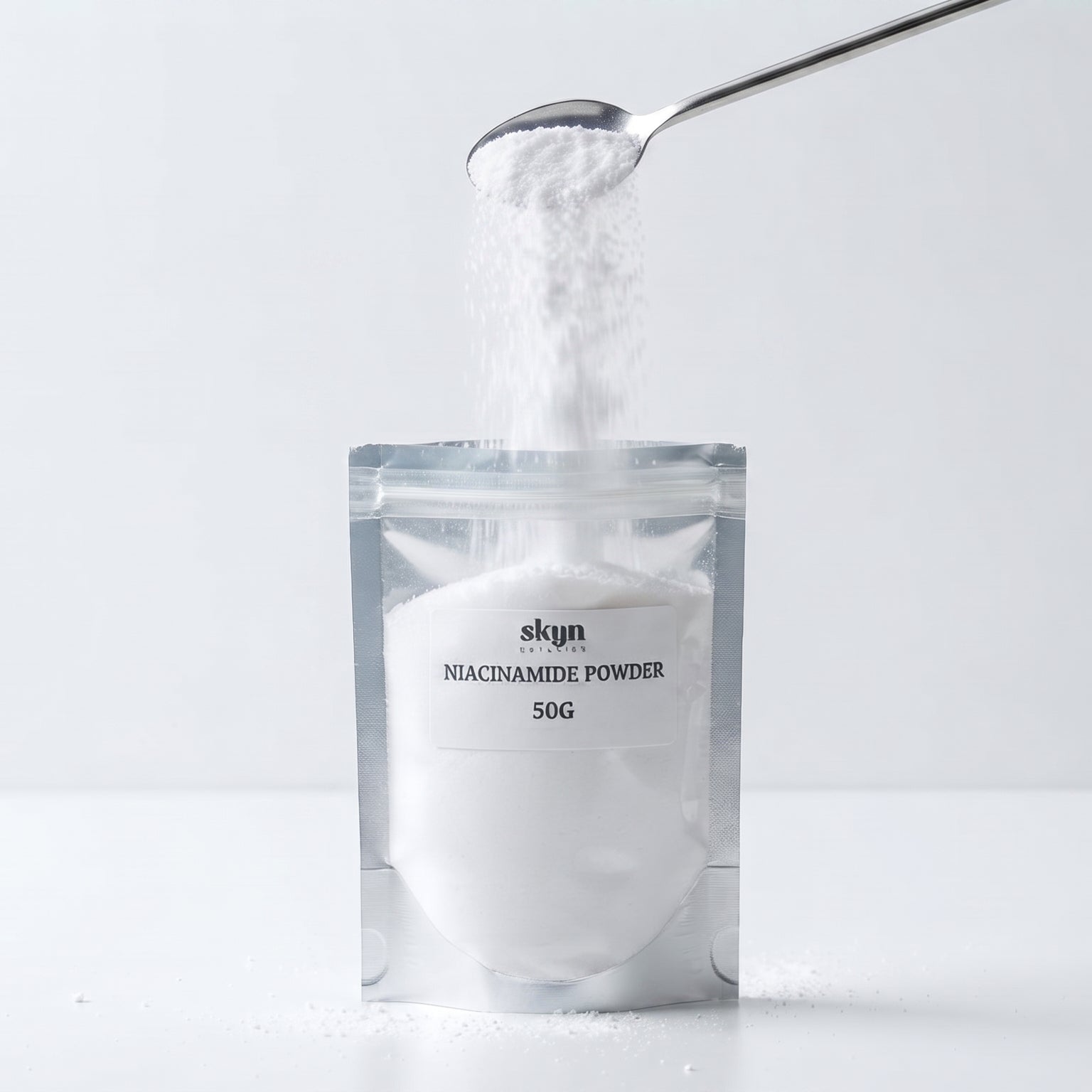 A silver stand-up pouch labeled 'skyn NIACINAMIDE POWDER 50G' on a white surface, with white powder being poured into it from a metal spoon against a light gray background.