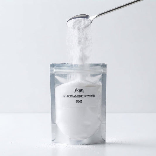 A silver stand-up pouch labeled 'skyn NIACINAMIDE POWDER 50G' on a white surface, with white powder being poured into it from a metal spoon against a light gray background.