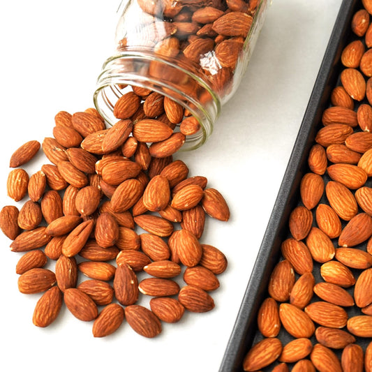 Almonds spilling from a jar onto a white surface with a tray of almonds nearby.