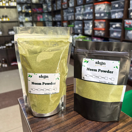 Two stand-up pouches of skjn Neem Powder displayed on a wooden counter in a retail store setting. One pouch is filled with light green powder and the other with dark green powder, both featuring white labels with the brand name 'skjn'.