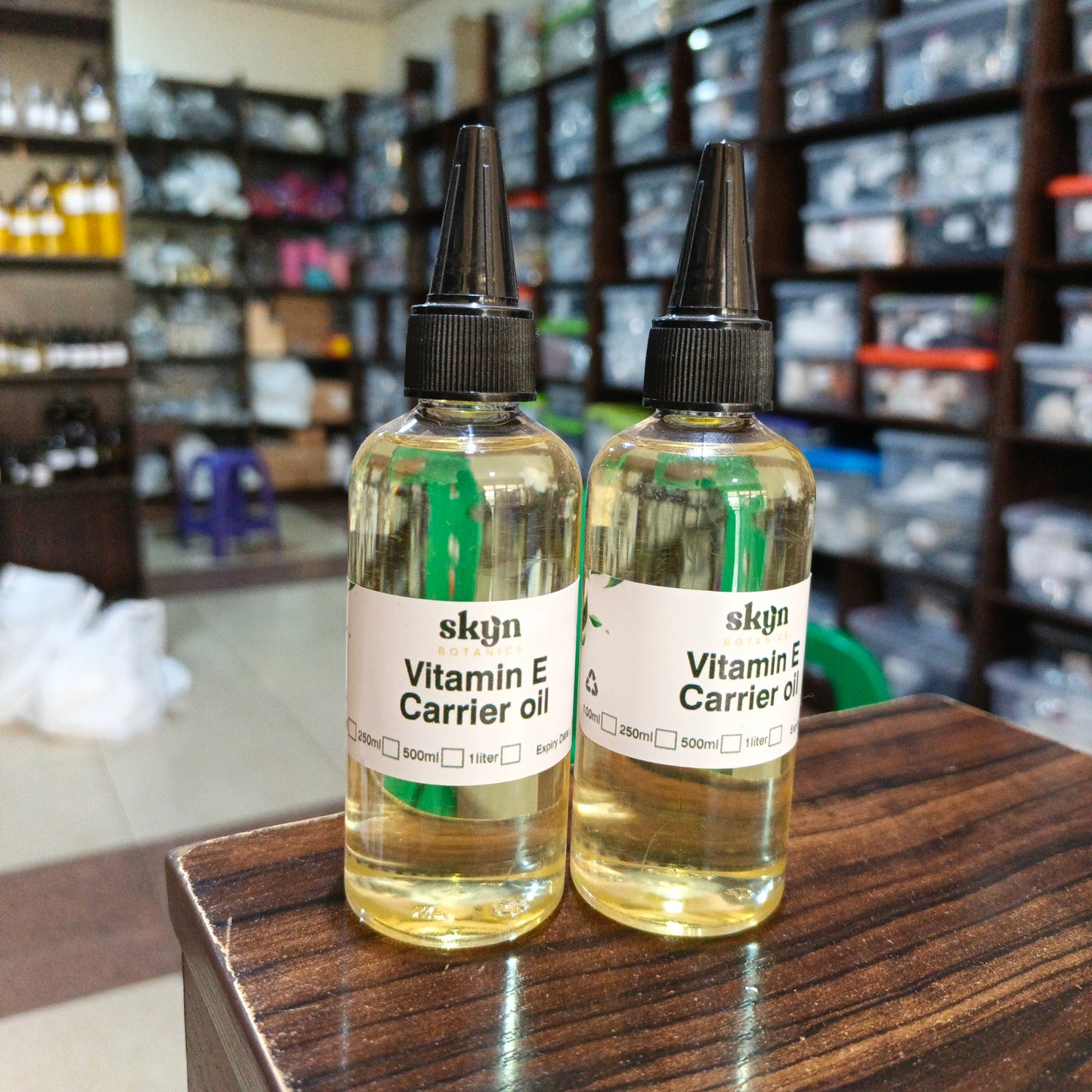 Two bottles of skqn Vitamin E Carrier oil on a wooden surface with shelves in the background.